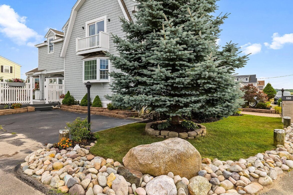 A two-story house with a well-maintained lawn, a large rock centerpiece, and a coniferous tree.