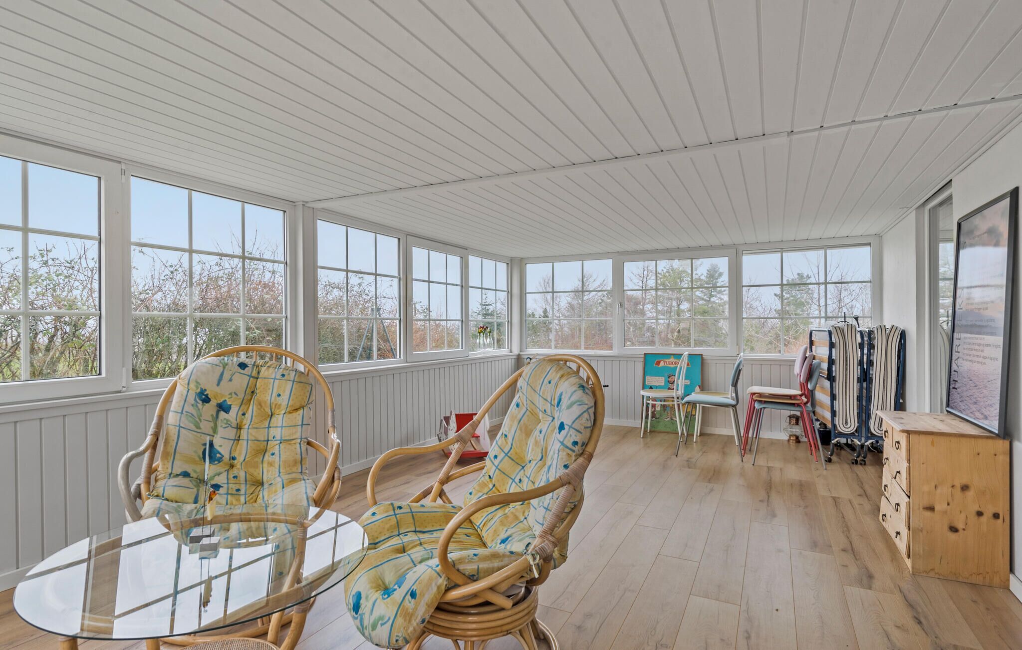 A sunroom with wicker chairs, a glass table, and a view of trees through large windows.