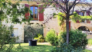 Ein traditionelles Steinhaus mit einem Vorgarten, der einen großen Baum mit violetten Blüten, einen gepflegten Rasen und eine Steinmauer zeigt.