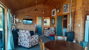 A cozy cabin interior with a wooden ceiling, a round dining table, two sofas, a TV, and a fireplace.