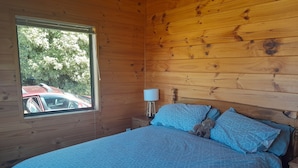 A wooden cabin room with a bed, a nightstand, a lamp, and a window with a view of a red car and trees.