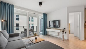 A modern living room with a grey sofa, a coffee table, a TV on a white cabinet, and a balcony with blue curtains. - Fewo 3-13,Ostsee-Nordmole (Olpenitz)
