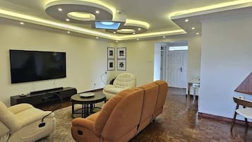 A modern living room with a flat-screen TV, a beige sofa, a brown recliner, and a wooden parquet floor.