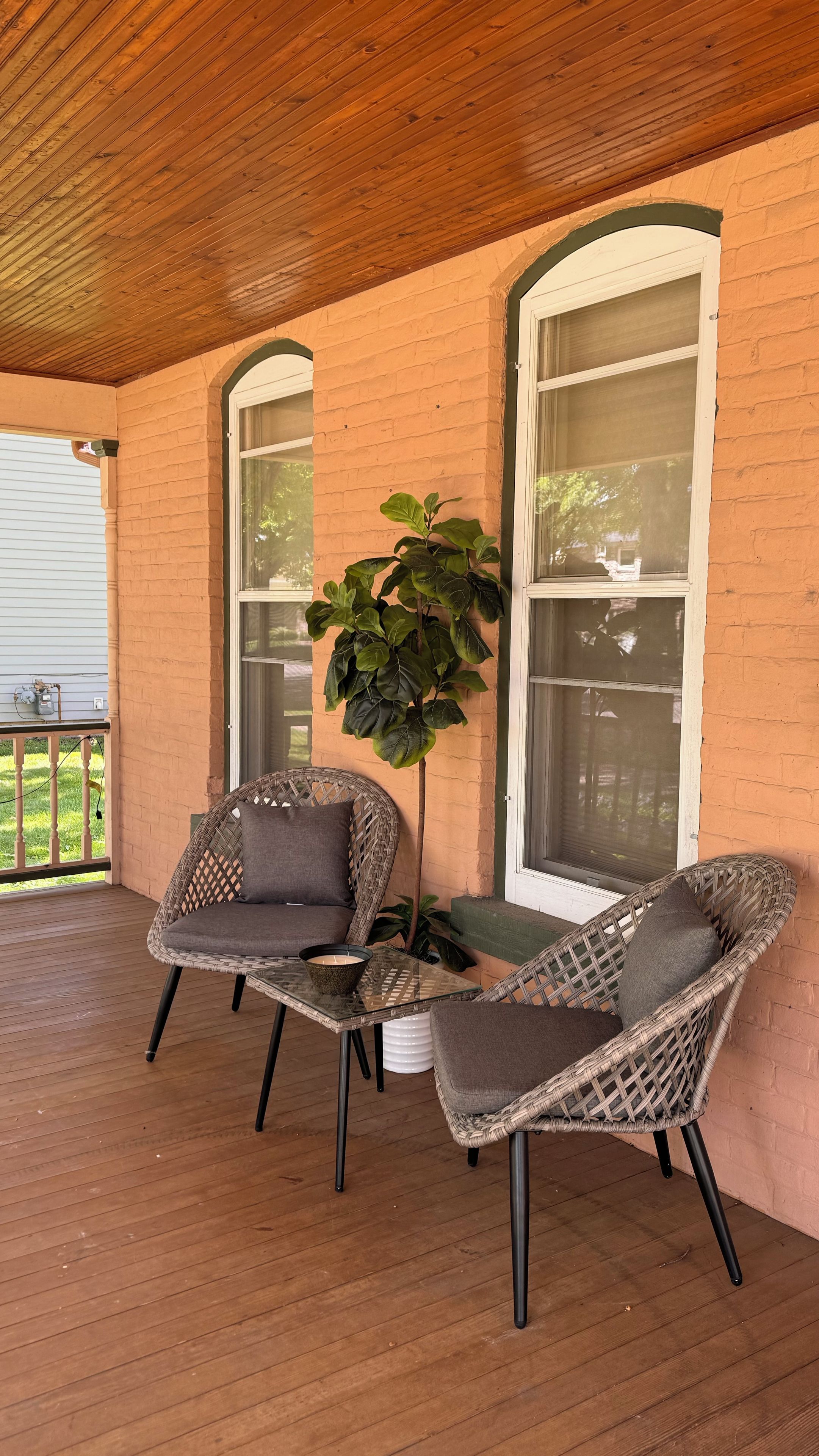 A covered porch with two wicker chairs and a table, a potted plant, and a brick wall with large windows.