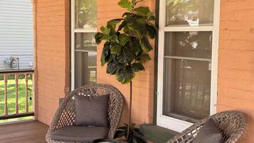 A covered porch with two wicker chairs and a table, a potted plant, and a brick wall with large windows.