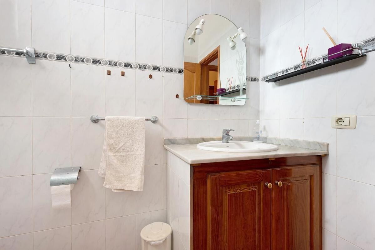 A bathroom with white tiles, a wooden vanity, a sink, a mirror, a towel rack, a toilet, and a shelf with toiletries.