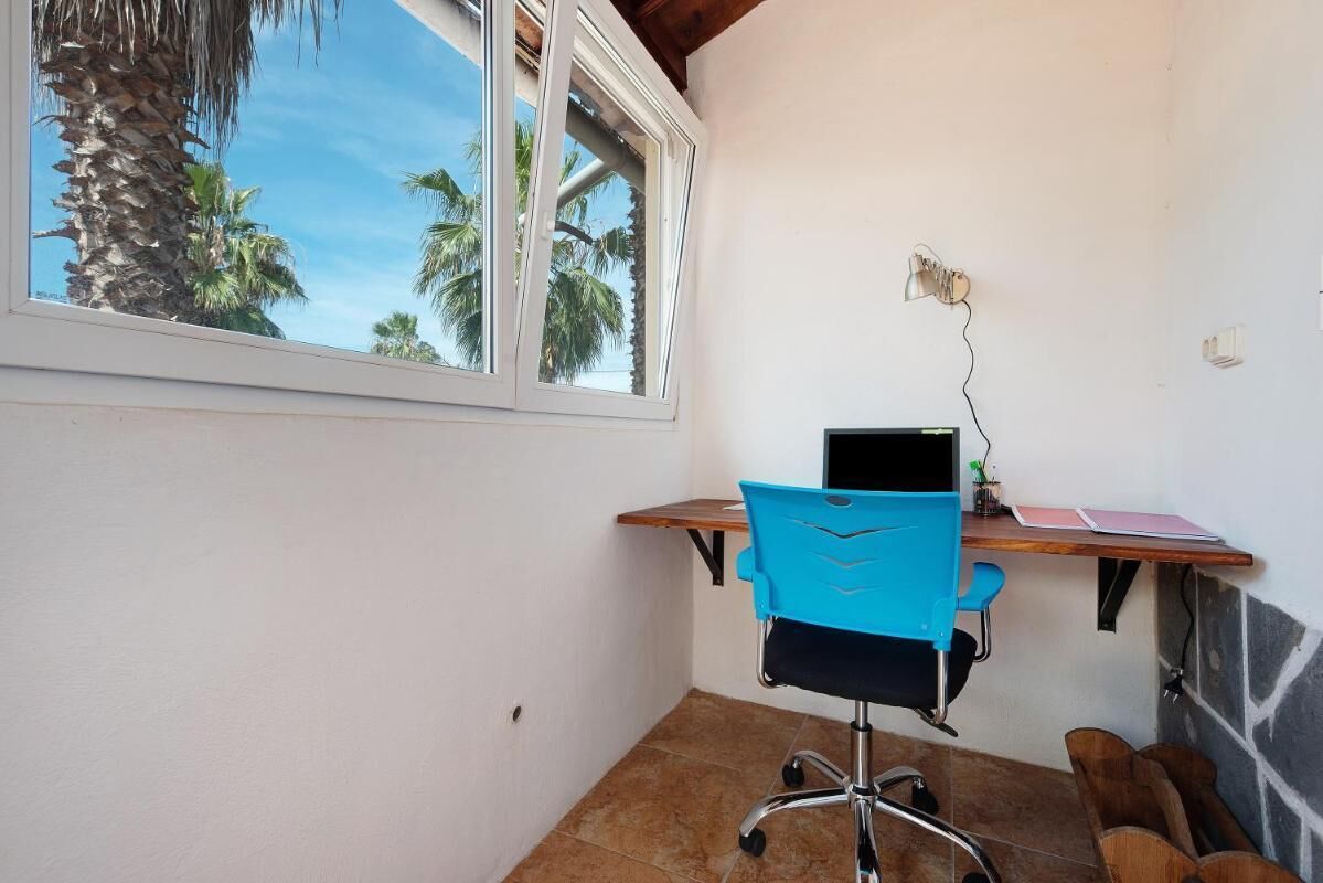 A small office space with a desk, a blue chair, and a laptop. There is a window with a view of palm trees.