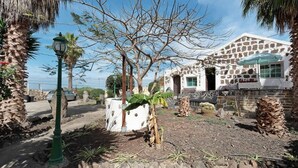 A stone building with a white facade and a green umbrella. There is a palm tree and a lamp post in the foreground. - Room-casa de Vacaciones El Cardón B2 (Buenavista del Norte)