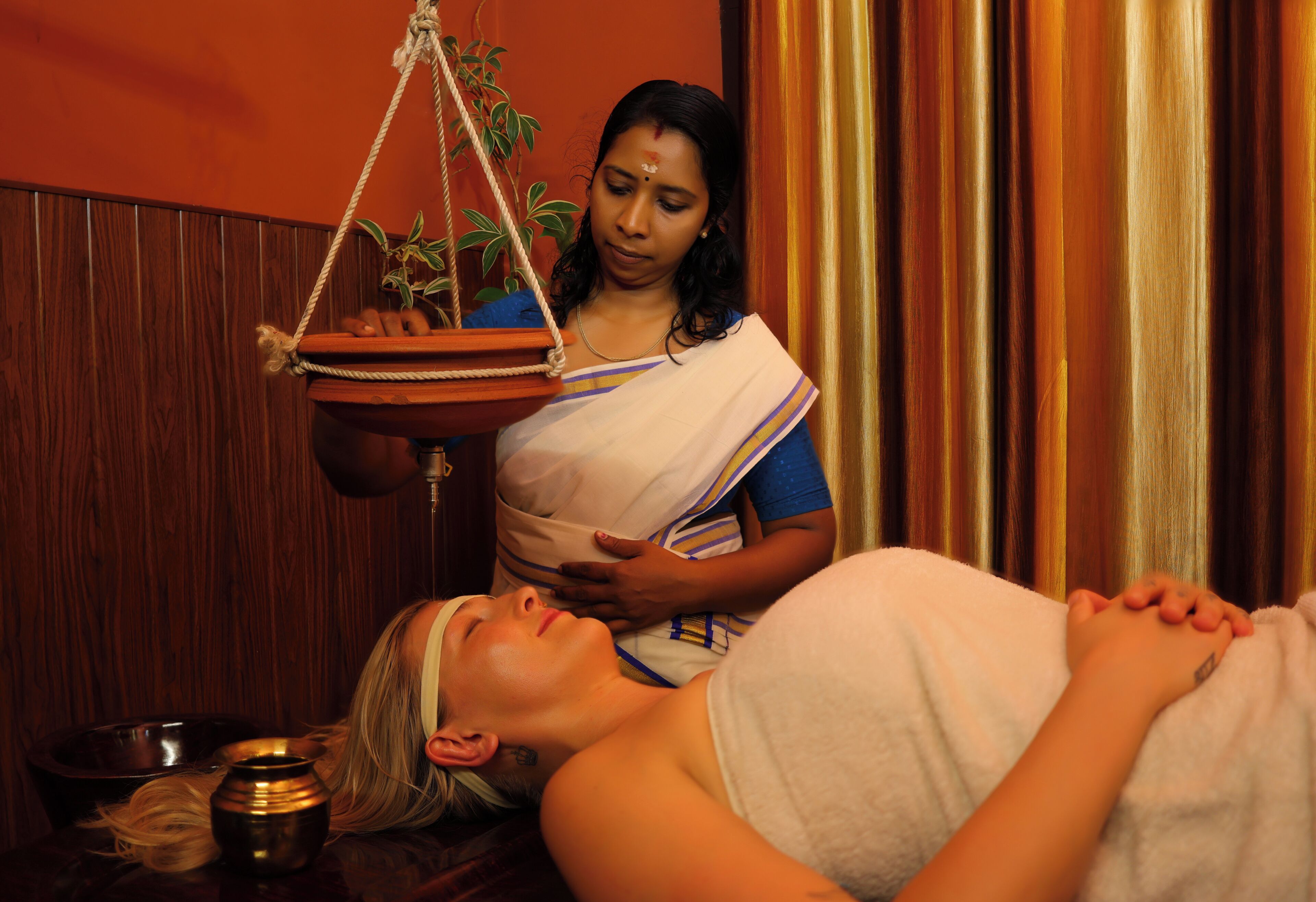 A person lying down with a woman standing beside them, holding a tray with a plant.