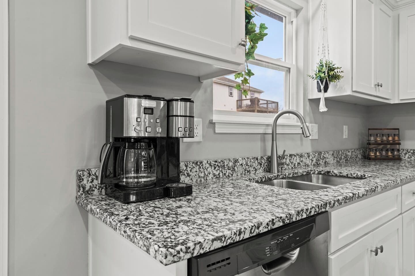 Una cocina con armarios blancos, encimera de granito, cafetera, lavavajillas y una ventana con una planta.