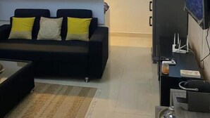Een woonkamer met een zwarte bank, gele kussens, een televisie en een keuken met kasten en een airconditioner.