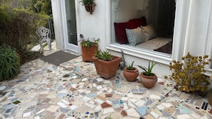 A white building with a mosaic-tiled patio, potted plants, and a window with a red cushion.