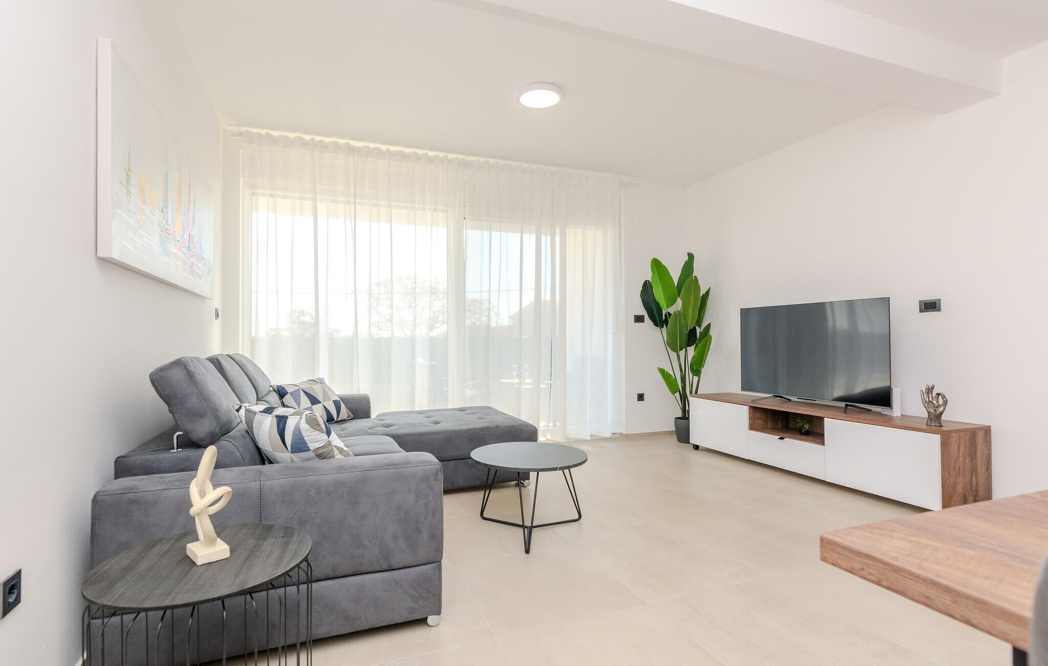 Uma sala de estar moderna com um sofá cinza, uma mesinha redonda, uma TV sobre um suporte de madeira e uma planta grande.