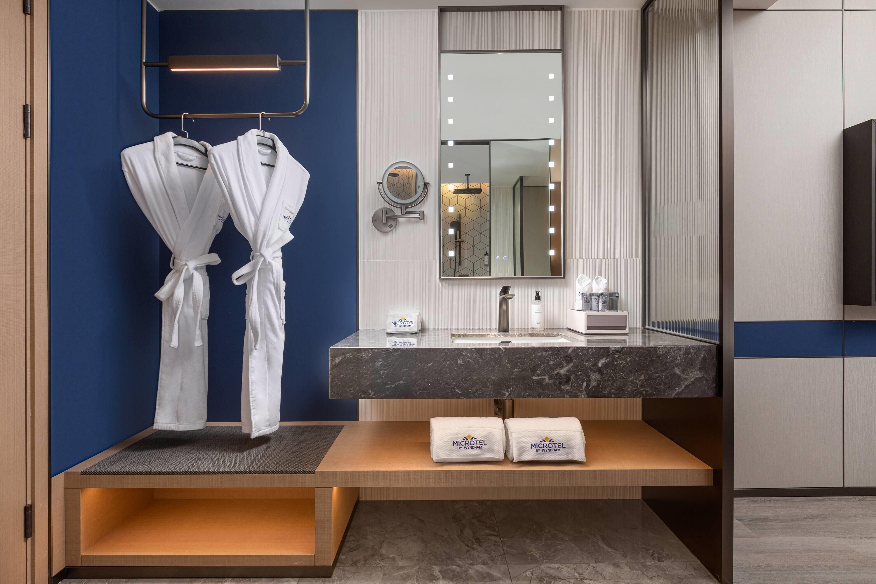 A modern bathroom with a blue accent wall, a vanity with a mirror, and two white robes hanging on a rack.