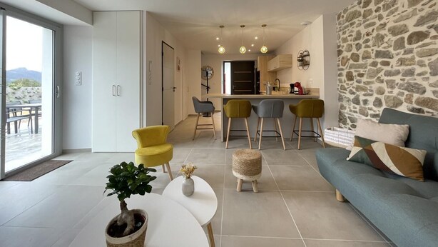 Un salon moderne avec un mur en pierre, un canapé, un coin repas avec des tabourets de bar et une cuisine en arrière-plan.
