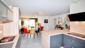 Eine moderne Küche mit einer zentralen Kochinsel, einem Essbereich mit bunten Stühlen und Blick zum Wohnzimmer.