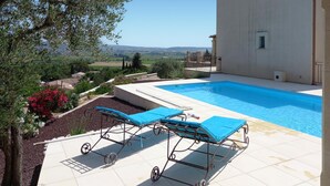 En swimmingpool med två solstolar, utsikt över landskapet och en byggnad med ett tak täckt av tegelpannor.