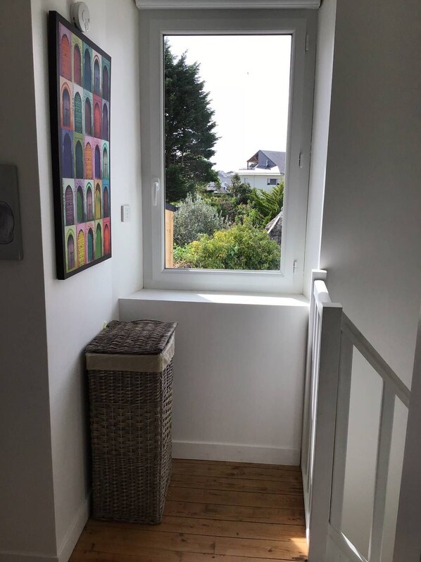Un couloir étroit avec une fenêtre, un panier en osier et une œuvre d’art colorée accrochée au mur.