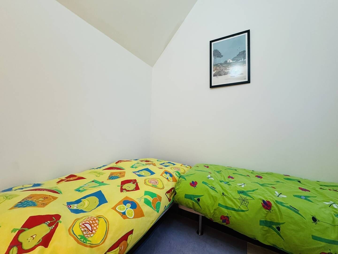 A small room with two single beds, one with a yellow and red patterned bedspread and the other with a green and red patterned bedspread. There is a framed picture on the wall above the beds.