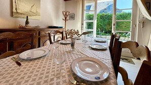 A dining room with a table set for four, wooden chairs, a chandelier, and a view of greenery through large windows.
