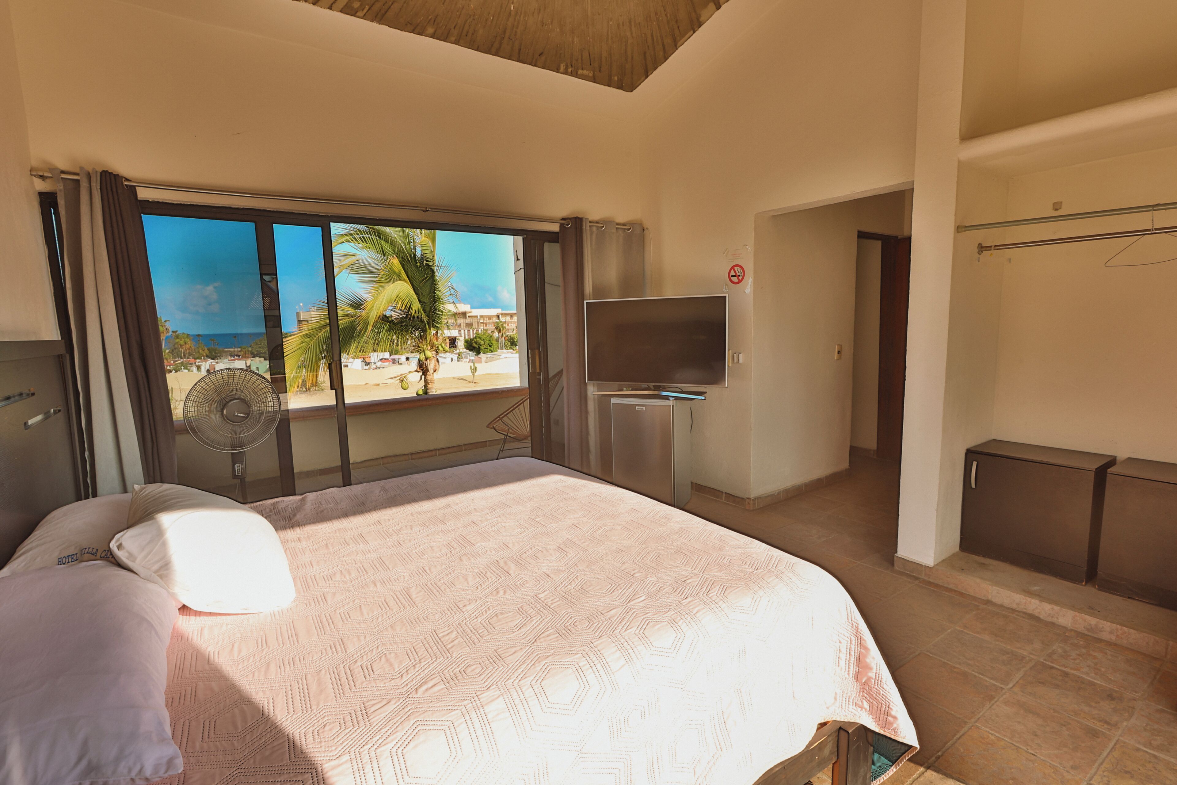 A hotel room with a bed, a TV, a balcony with a view of palm trees and buildings, and a door leading to another room.