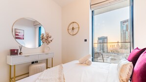 A modern hotel room with a large bed, a round mirror, a bedside table with a vase, and a view of the city through a large window. - City Views 1BR Close to Golf Course & Attractions, Binghatti Venus, Jumeirah Village Circle (Dubai)