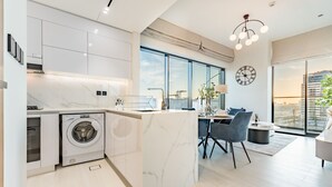 A modern kitchen with a built-in washing machine, a dining area with a table and chairs, and large windows offering a view of the city. - City Views 1BR Close to Golf Course & Attractions, Binghatti Venus, Jumeirah Village Circle (Dubai)