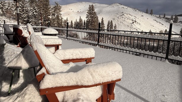 Sillones de exterior cubiertos de nieve, una valla de metal y un paisaje montañoso nevado.