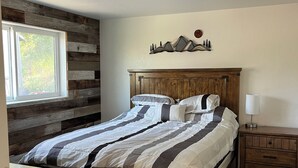 Un dormitorio con una cama de madera, una mesita de noche, un estante en la pared con adornos y una ventana con vistas a la vegetación.