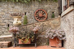 A stone wall with a rustic wooden wheel, potted plants, and a small window with shutters.