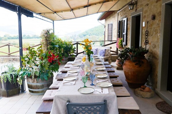 An outdoor dining area with a long table set for a meal, surrounded by potted plants and a stone building.