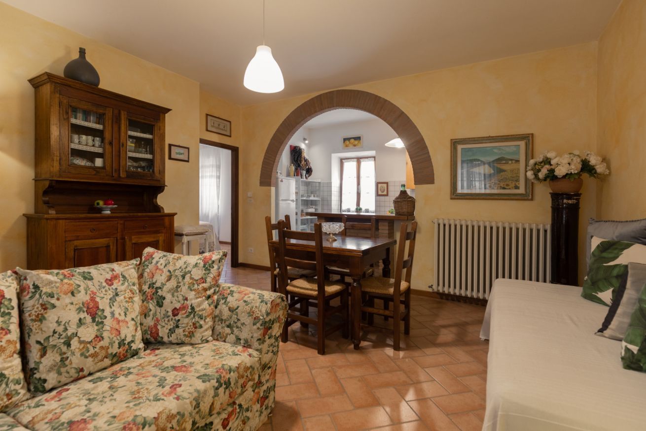 A dining area with a wooden table and chairs, a floral sofa, and a kitchen visible through an archway.