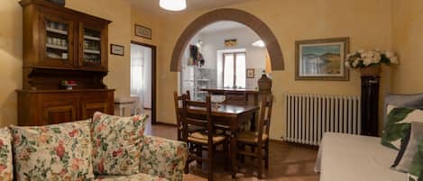 A dining area with a wooden table and chairs, a floral sofa, and a kitchen visible through an archway.