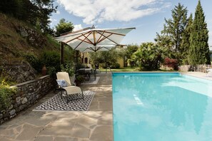 Une piscine avec une terrasse en pierre, du mobilier d’extérieur et un grand parasol.