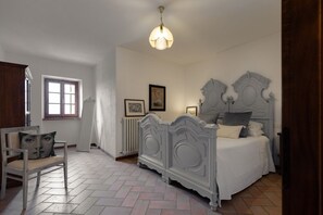 Un dormitorio con una cama, una silla, un radiador, una ventana y una lámpara en el techo.