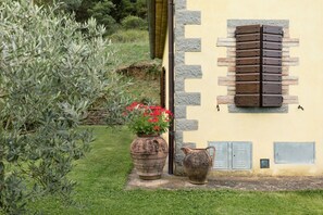 A building with a stone wall, a wooden shutter window, a potted plant with red flowers, and a weathered pot. - Villa Essiccatoio by MMega (Lisciano Niccone)
