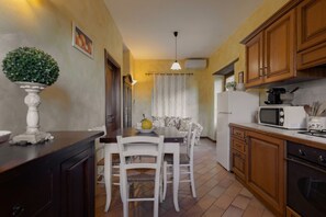 A compact kitchen with wooden cabinets, a dining table with chairs, a white refrigerator, and a microwave. - Villa Essiccatoio by MMega (Lisciano Niccone)