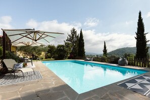 A swimming pool with lounge chairs and umbrellas, surrounded by stone flooring and tall trees.
