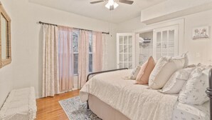 Ein Schlafzimmer mit einem Bett, einem Deckenventilator, einem Fenster mit Vorhängen, einem Nachttisch und einer Tür zu einem anderen Raum.