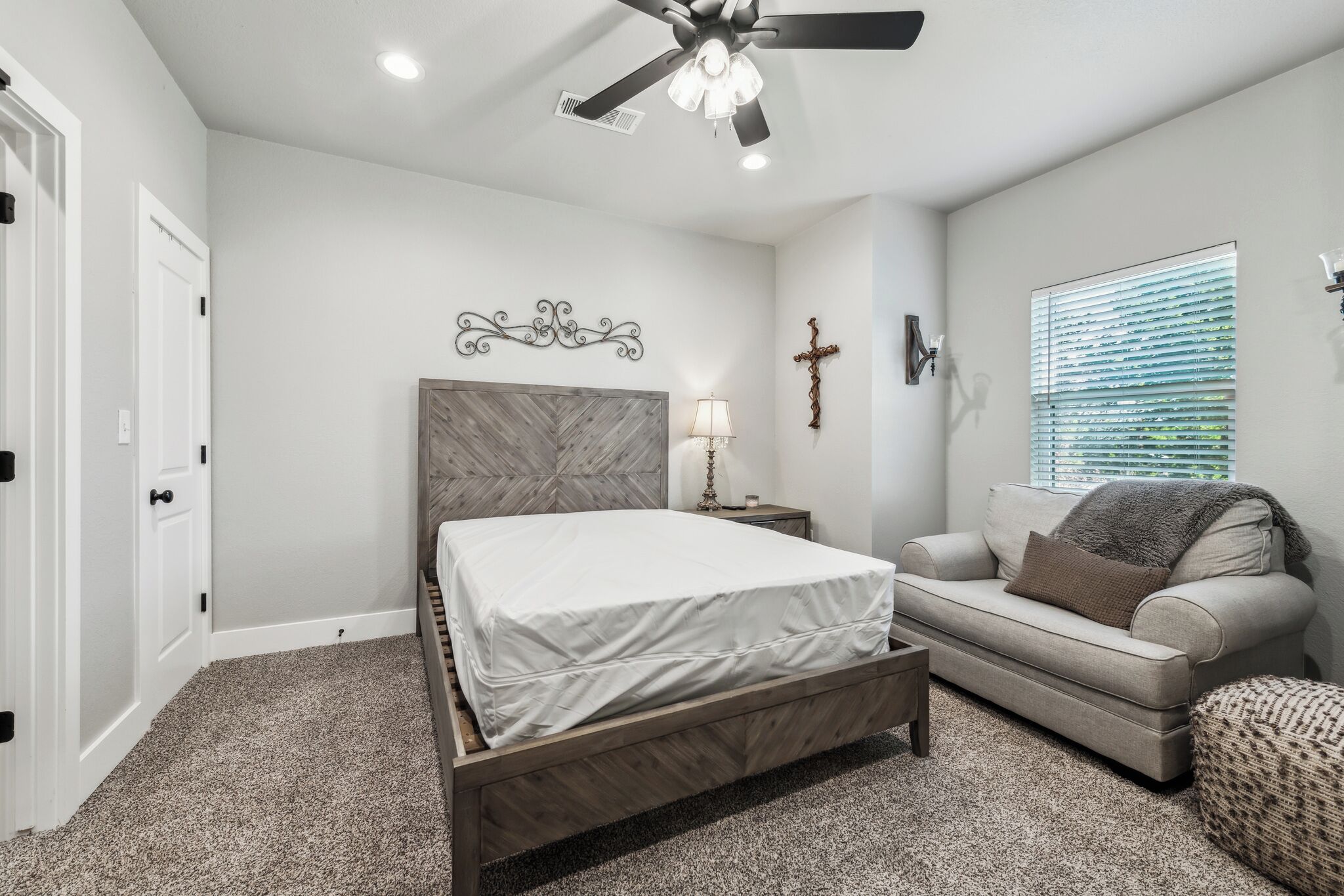 A bedroom with a bed, a nightstand, a sofa, a ceiling fan, a cross on the wall, and a window with blinds.