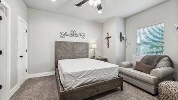 A bedroom with a bed, a nightstand, a sofa, a ceiling fan, a cross on the wall, and a window with blinds.