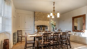 A dining room with a large wooden table set for six, surrounded by wooden chairs. A chandelier hangs from the ceiling, and a stone fireplace is visible in the background.
