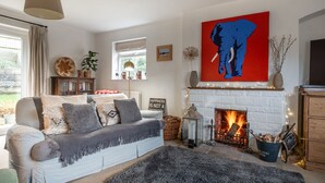 A cozy living room with a white fireplace, a sofa with patterned cushions, a large abstract painting, and a view of the outdoors.