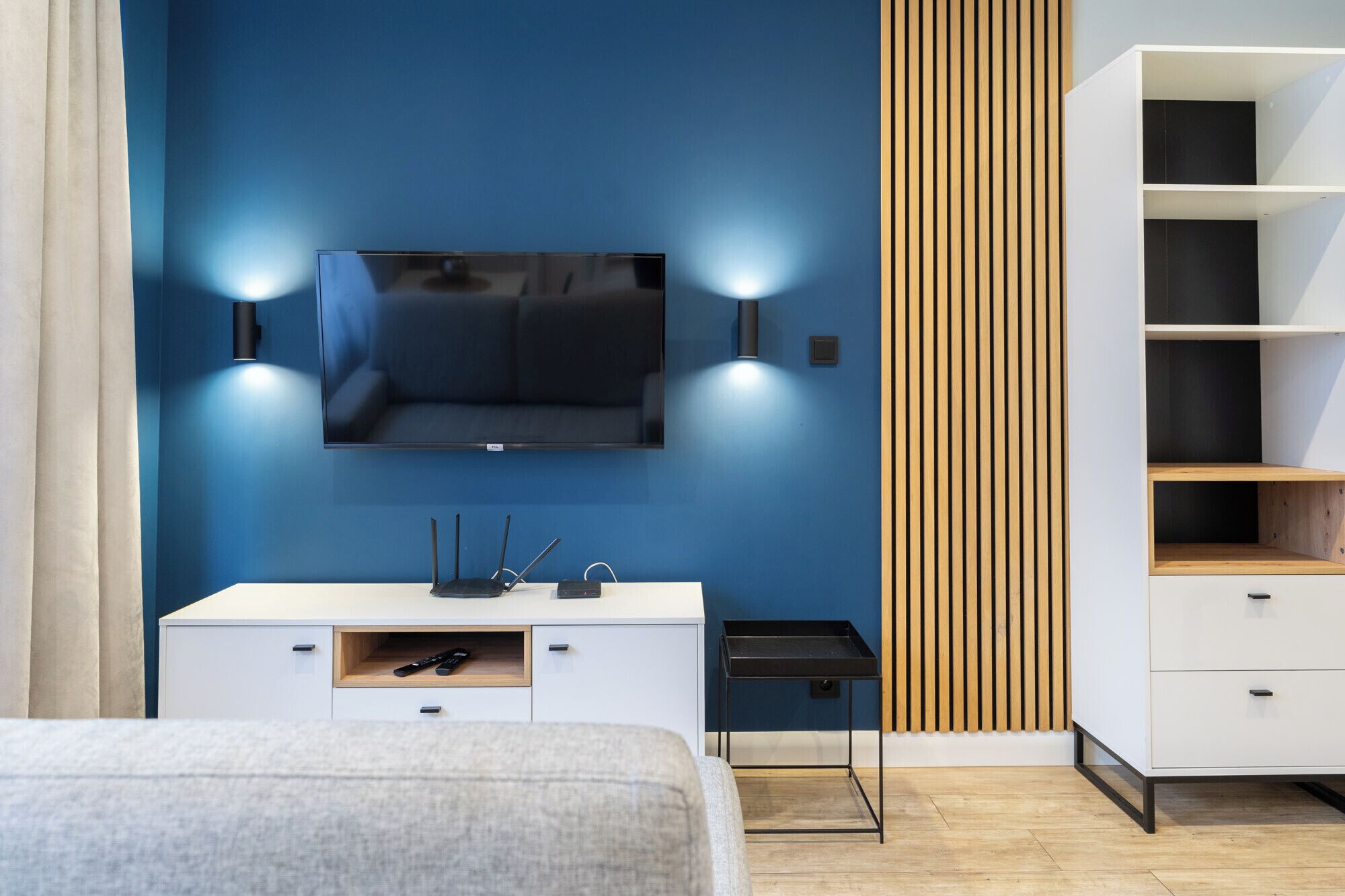 Camera moderna con una grande TV montata su una parete blu, un comodino bianco con cassetto, un pannello di legno a listelli e una libreria a muro.
