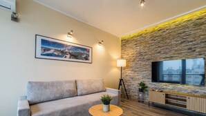 Una sala de estar moderna con un sofá, una mesa redonda de madera con una planta, un televisor montado y una pared de piedra.