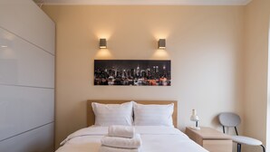 Una cama bien hecha con sábanas blancas y una toalla doblada, una mesita de noche con una lámpara, una silla y un cuadro de un paisaje urbano en la pared.