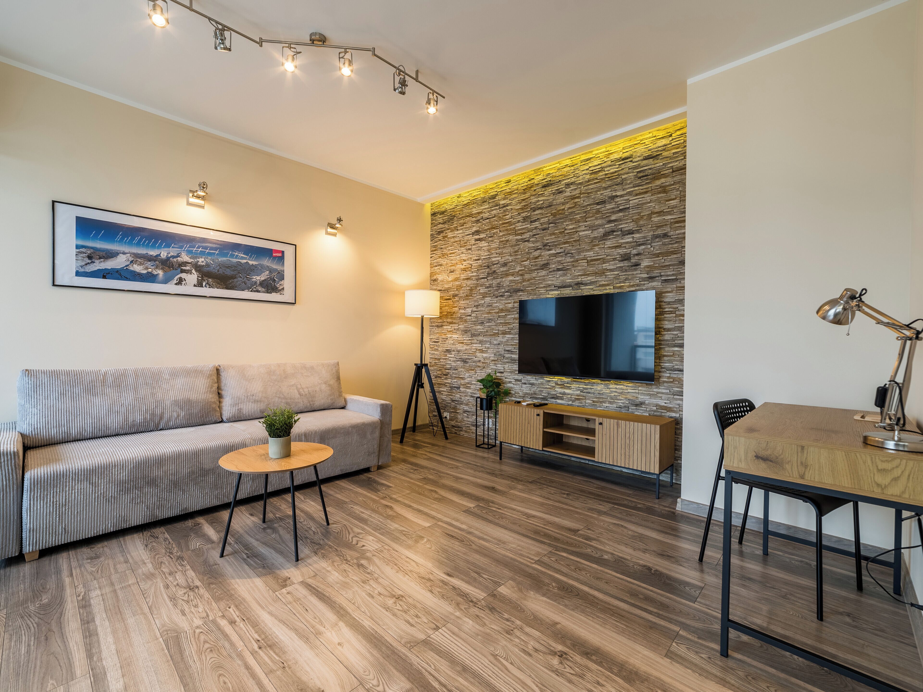 Een moderne woonkamer met een bank, een salontafel, een tv bevestigd aan een stenen muur, en een bureau met een lamp.