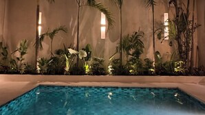 Una piscina nocturna con diseño de cuadros, rodeada de palmeras y plantas, con luces en la pared.
