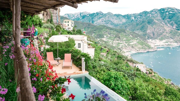 En pool med udsigt til havet og bjergene, omgivet af frodig vegetation og et træbeklædt pergola.