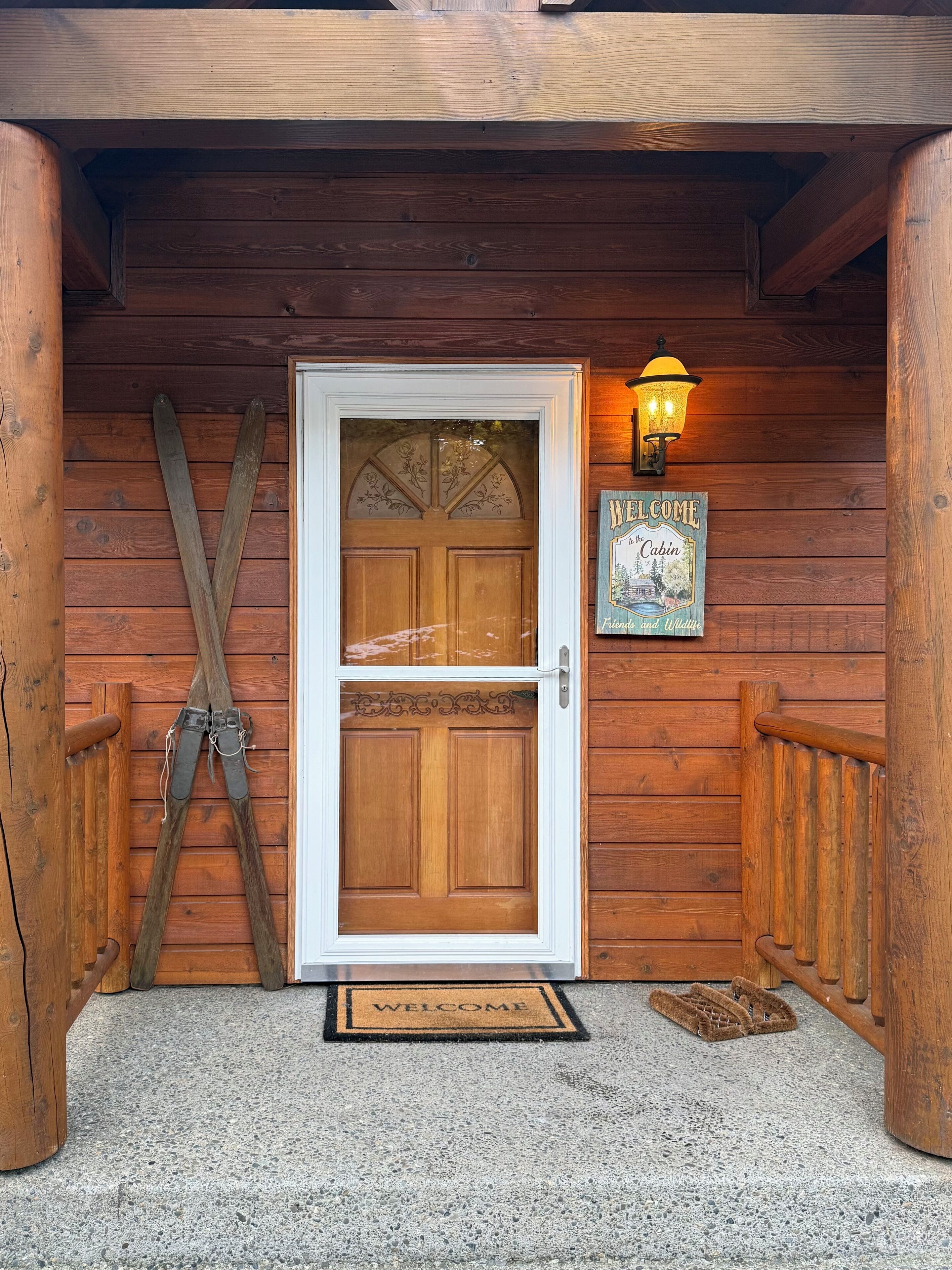Entrada a una cabaña de madera con puerta doble, felpudo y esquíes apoyados en la pared, además de un letrero de bienvenida.